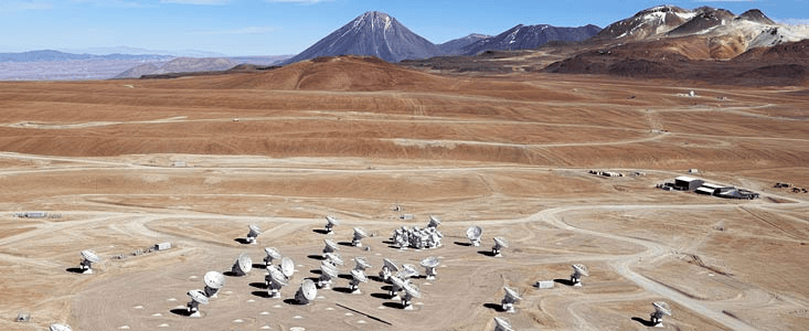Aerial view of a desert landscape featuring multiple satellite dishes, with mountains in the background.