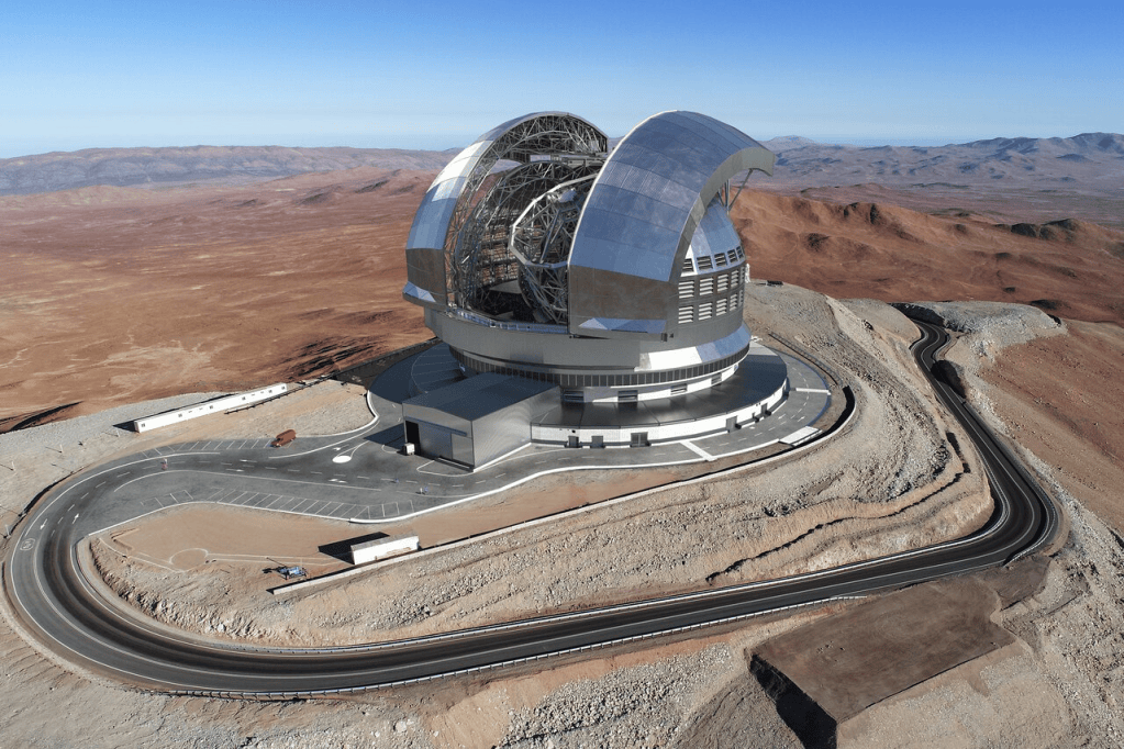 Aerial view of a modern astronomical observatory situated in a rugged, mountainous landscape under a clear blue sky.