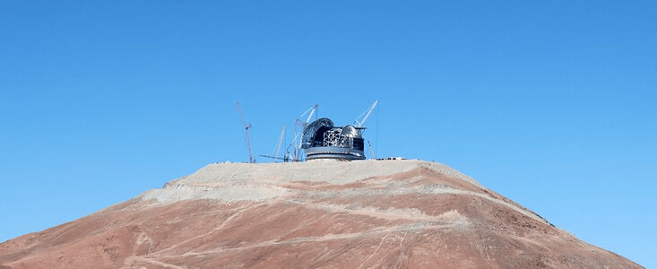 A large observatory structure atop a barren, reddish mountain, with clear blue skies in the background.