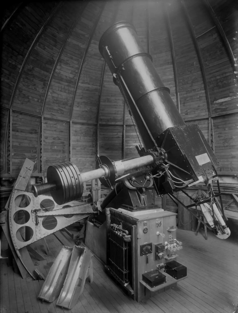 A vintage telescope positioned inside an observatory, showcasing its large metallic structure and various controls against a wooden interior.