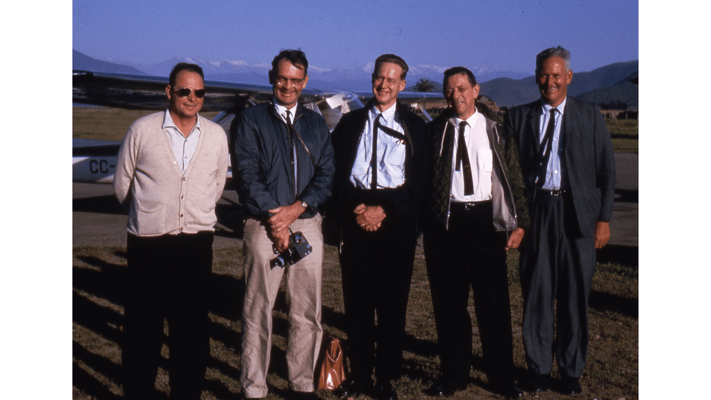 A group of five men standing together outdoors, with mountains visible in the background and an aircraft to the side. The men are dressed in various suits and casual attire.