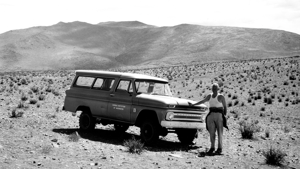 A black and white photo of a man standing next to a vintage Chevrolet Suburban truck in a desert landscape, with mountains in the background.