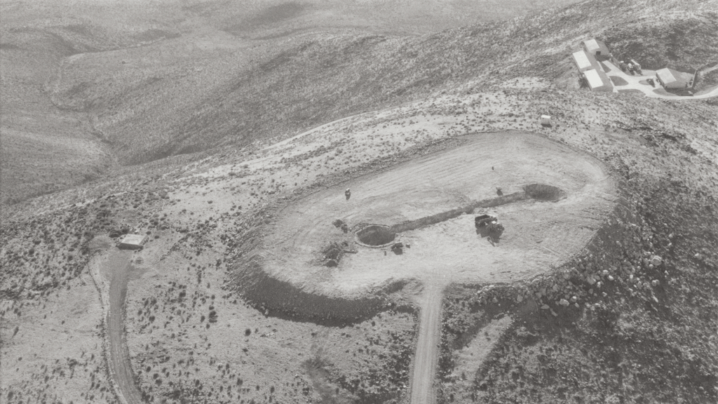 Aerial view of a barren landscape featuring an oval excavation site with two circular holes and machinery, surrounded by rocky terrain and sparse vegetation.