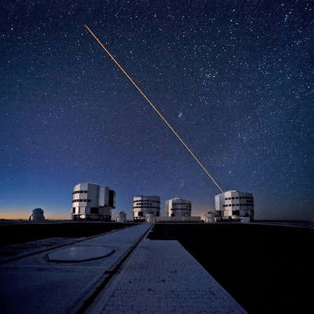A night sky filled with stars above a series of modern observatory buildings, with a bright laser beam extending upward into the cosmos.